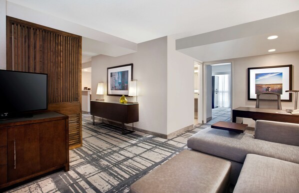 A modern hotel room with a flat-screen TV, a wooden accent wall, a sofa, a coffee table, and a desk with a lamp.