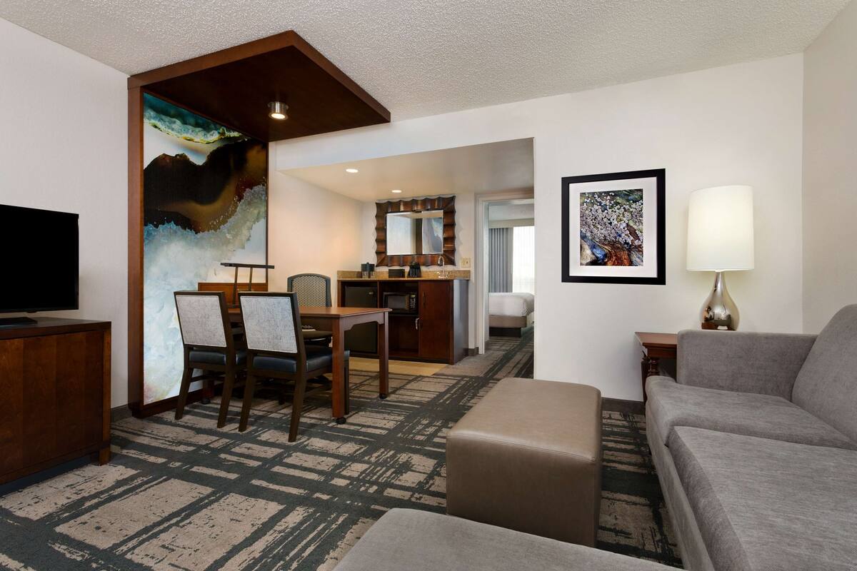 A hotel room with a living area, a dining table with chairs, a sofa, a television, a lamp, and artwork on the walls.