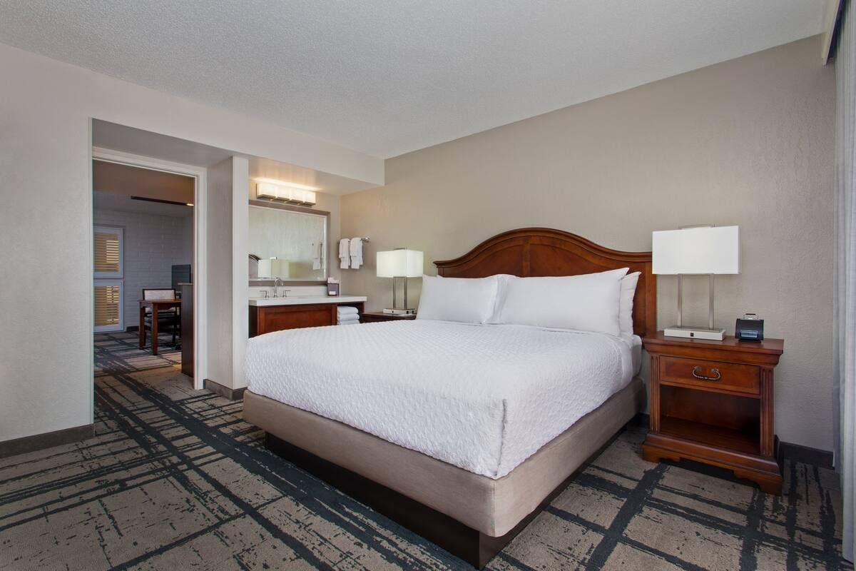 A hotel room with a large bed, bedside tables with lamps, a mirror, and a bathroom visible through an open door.