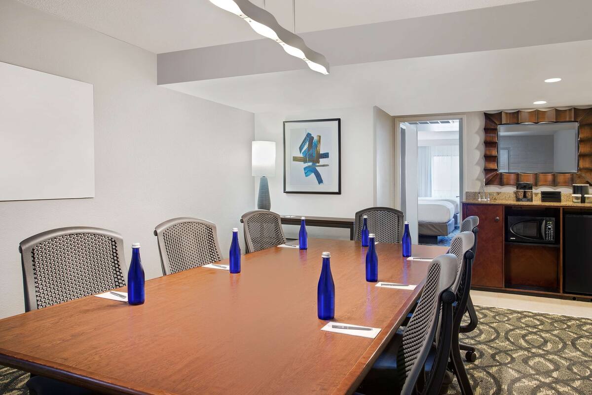 A conference room with a long table, six chairs, and blue water bottles. There is a microwave and a television on a counter.