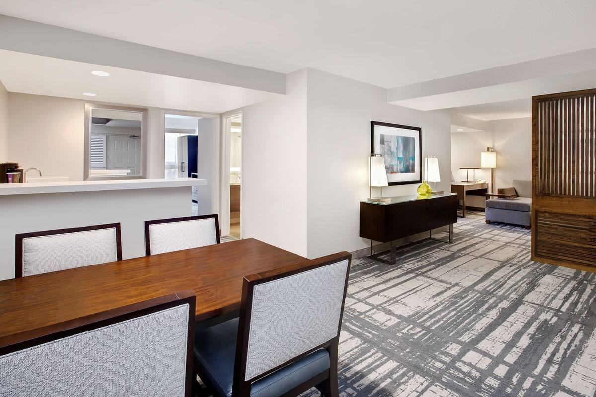 A hotel room with a dining area, a living area with a sofa and armchair, a wooden sideboard, and a large window.