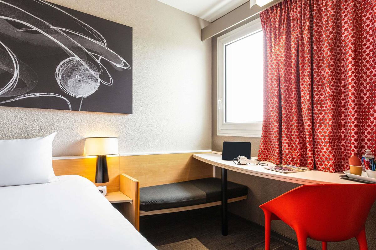 Una habitación de hotel con cama, escritorio con ordenador portátil, silla, lámpara y ventana con cortinas.