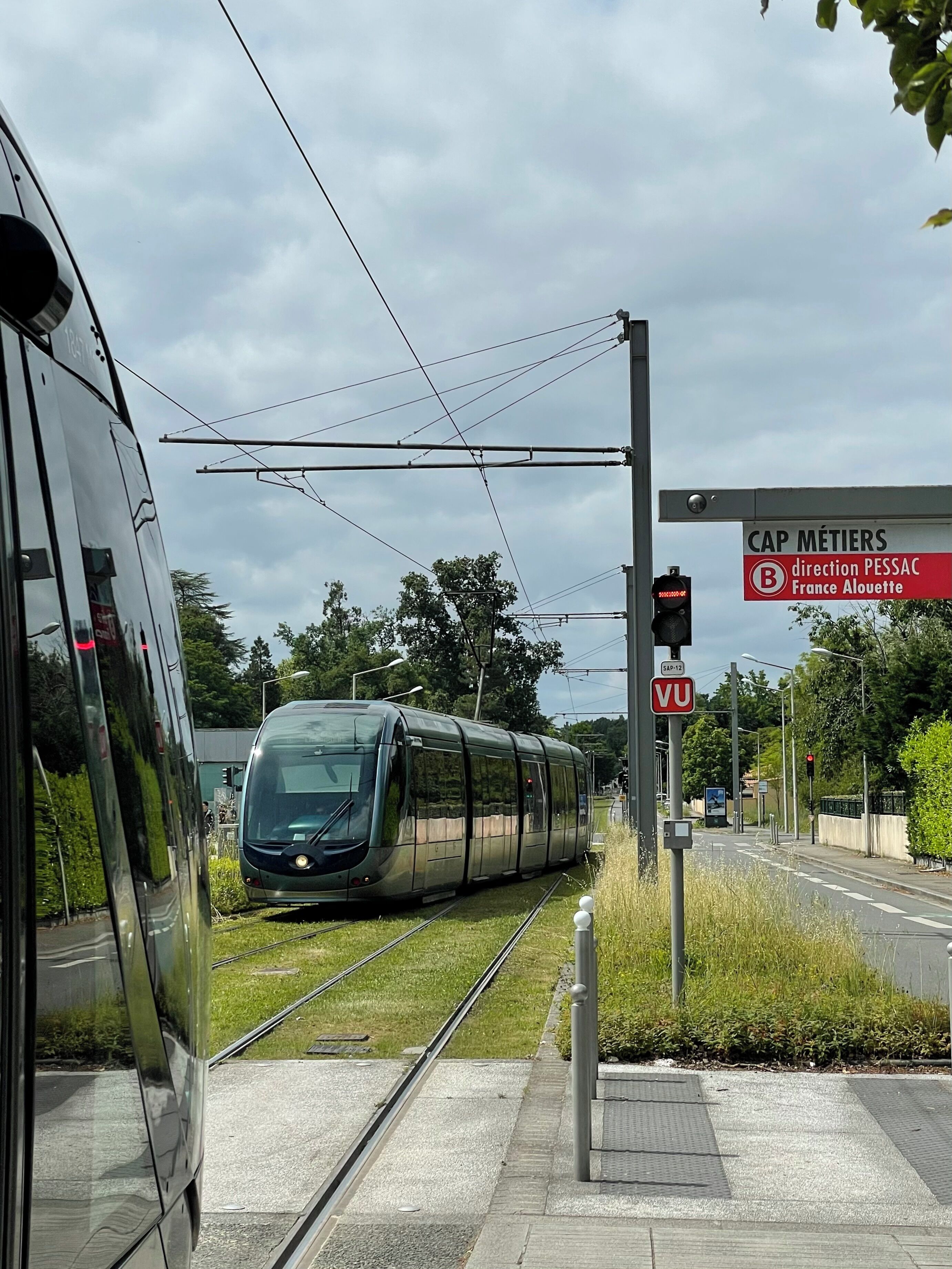 Een moderne tram bij een station met bovenleiding, een voetgangersoversteek en een bord waarop 'CAP MÉTIERS' en 'richting PESSAC, Frankrijk Alouette' staat.