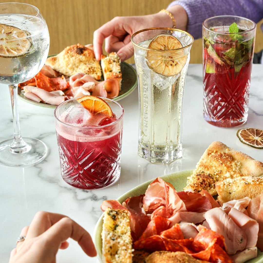 Una mesa con diversas bebidas, pan y embutidos cortados.