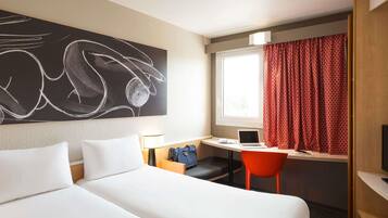 A hotel room with a bed, a desk with a laptop, a chair, a lamp, and a large abstract painting on the wall.