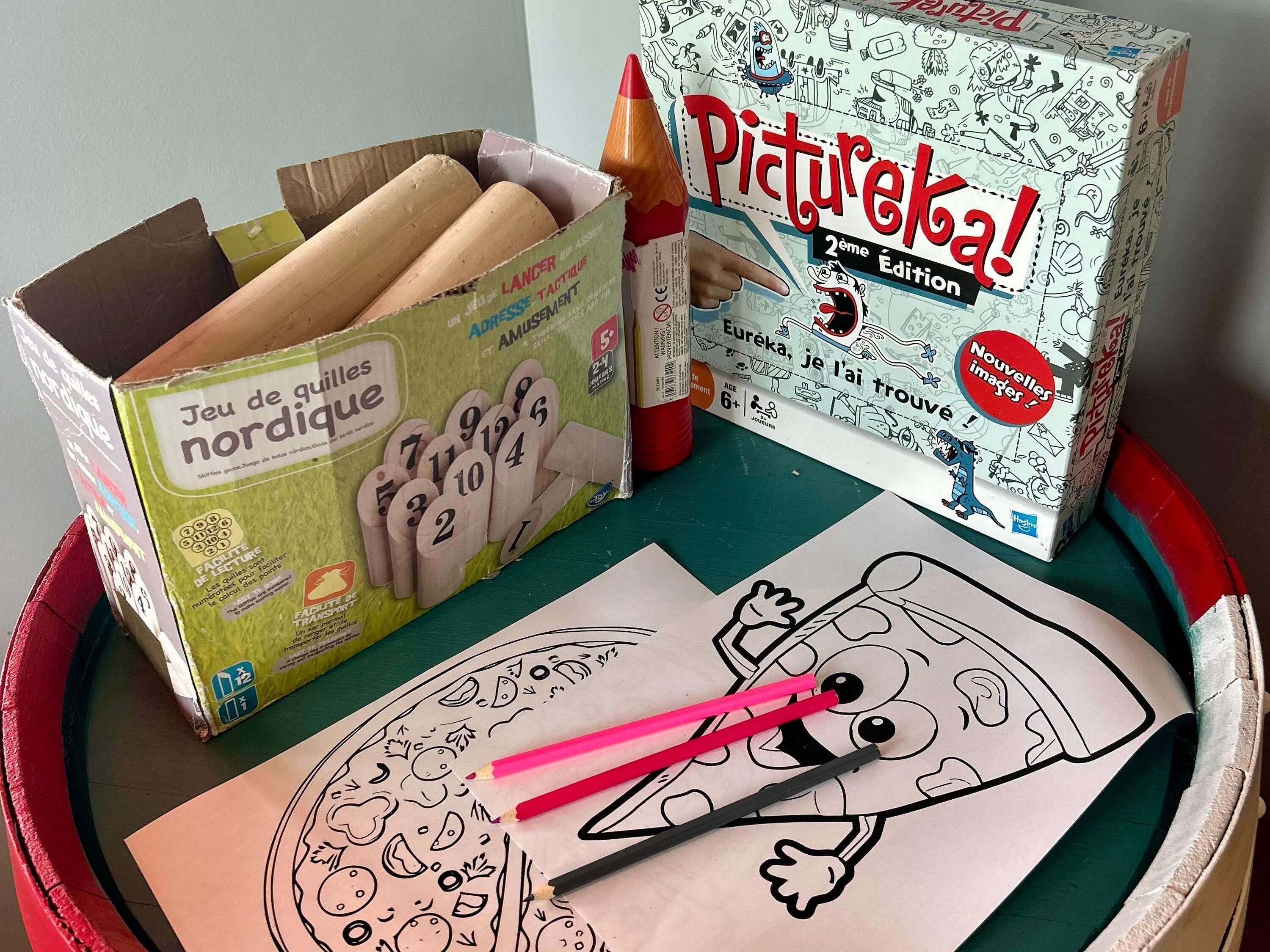 Une boîte de jeu étiquetée « Jeu de quilles nordique » et une boîte « Pictureleka ! 2e édition » sont posées sur une table, à côté d’un dessin et de crayons de couleur.