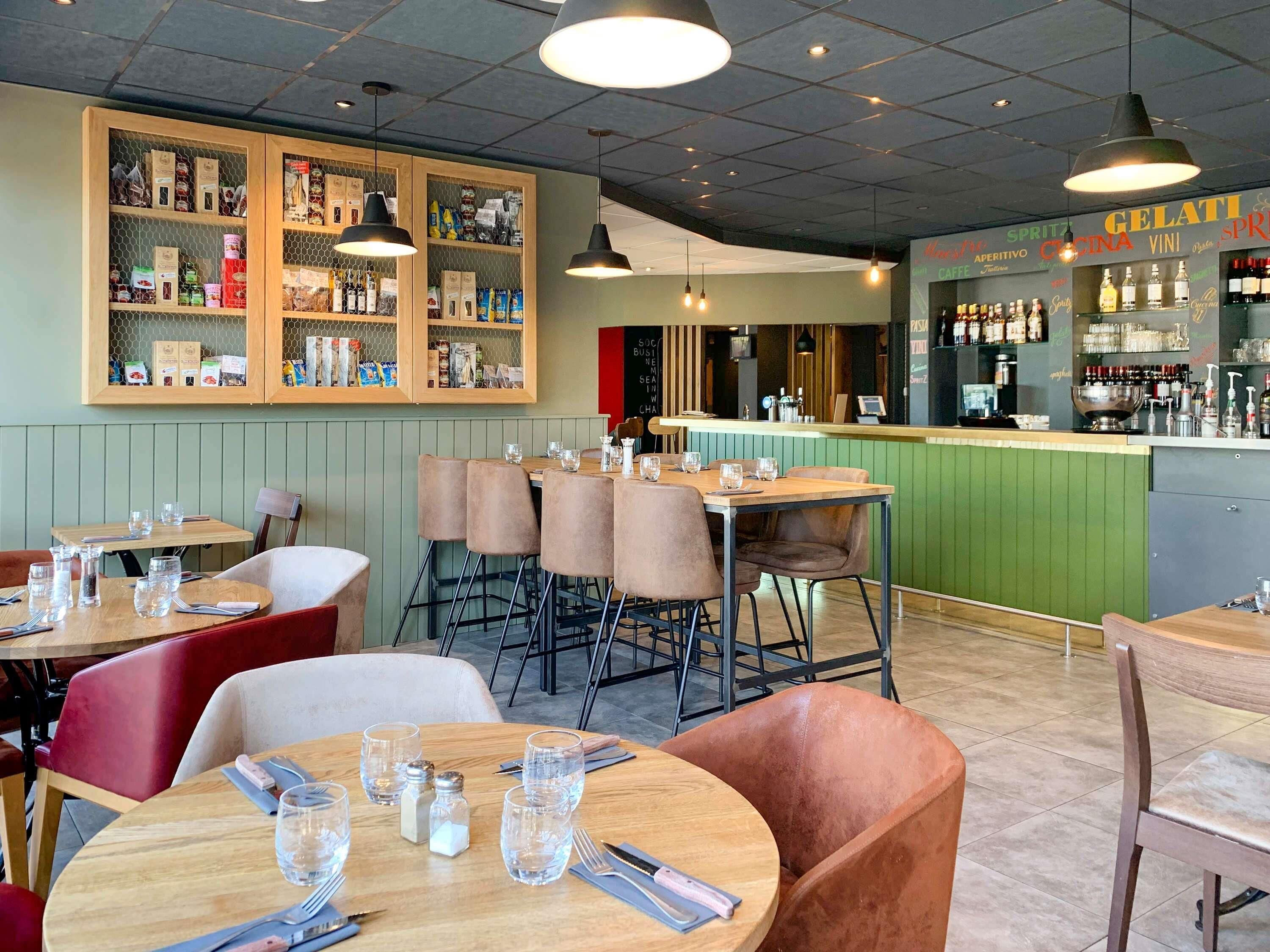 L’intérieur d’un restaurant présente de nombreuses tables et chaises, un bar et des étagères remplies de divers produits alimentaires et condiments.