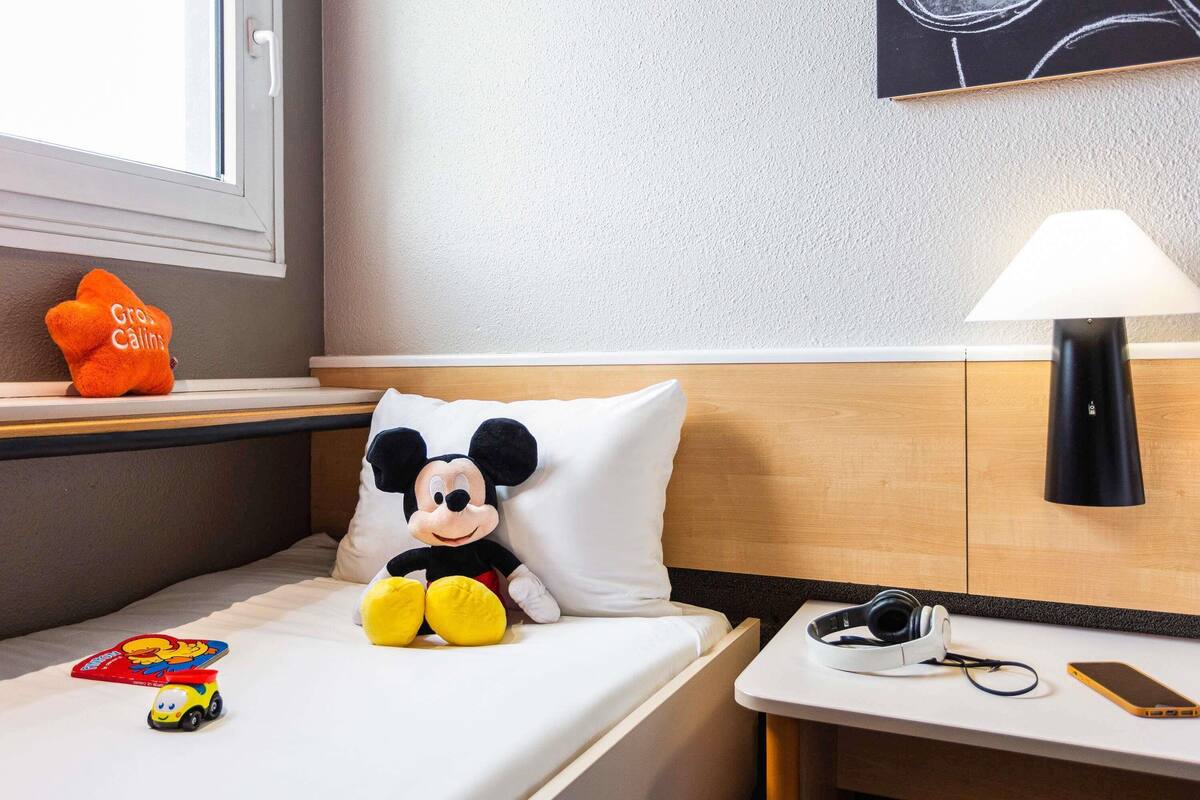 Una habitación de hotel con una cama, un cojín de Mickey Mouse, un pequeño coche de juguete, una lámpara, auriculares y un teléfono inteligente sobre una mesita de noche.