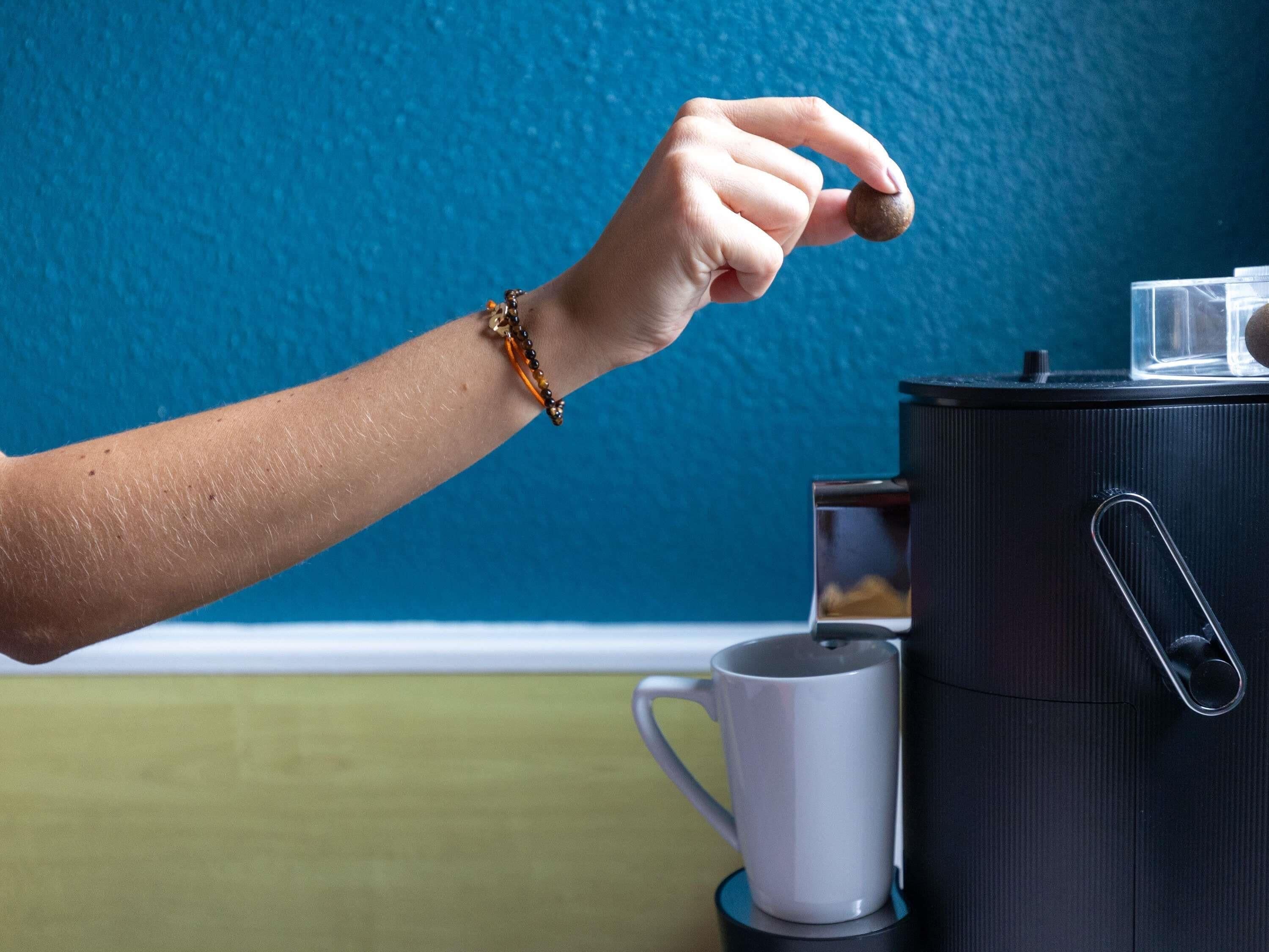 A hand holding a small object near a coffee maker and a mug on a table.