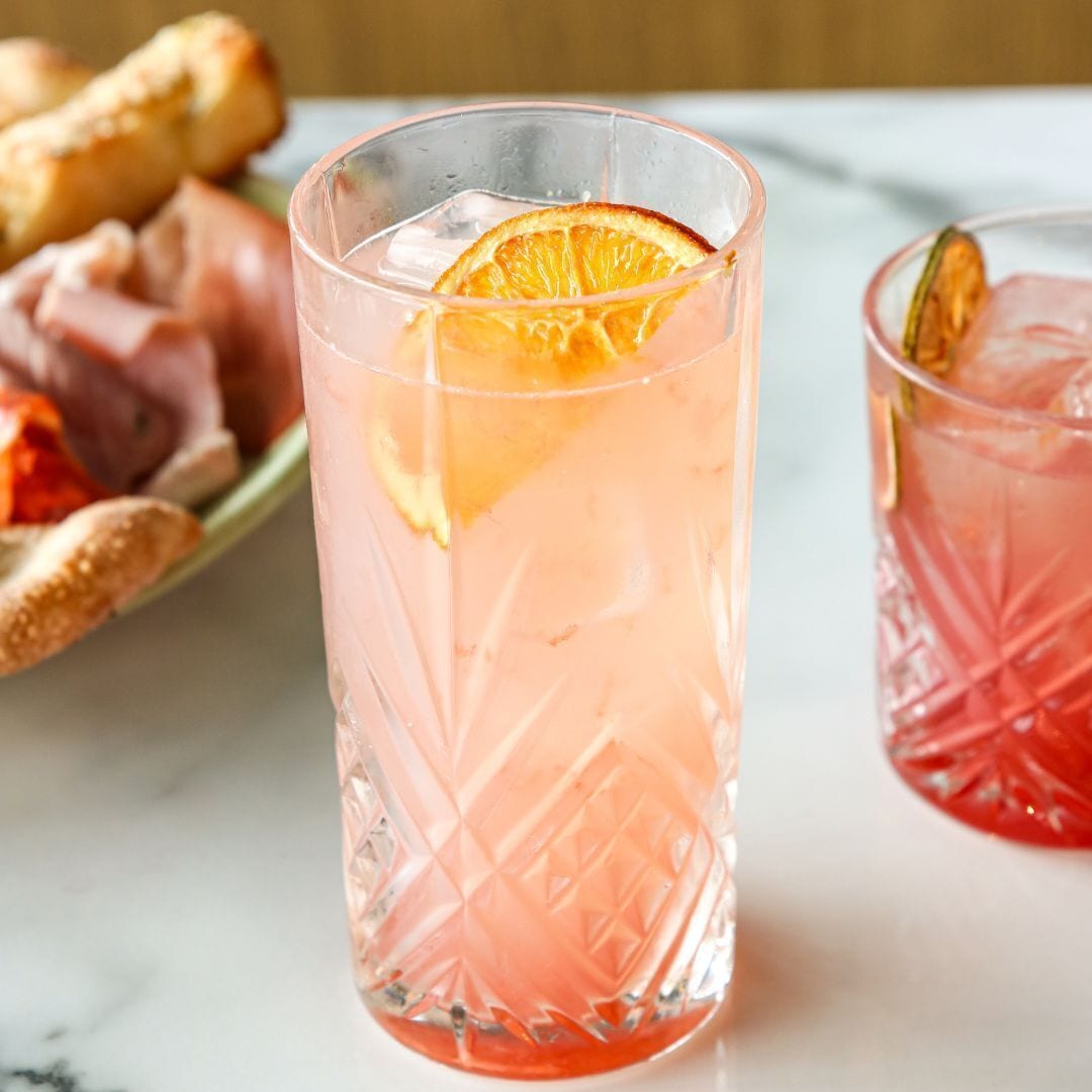 A glass of pink beverage with an orange slice, accompanied by a plate of food.