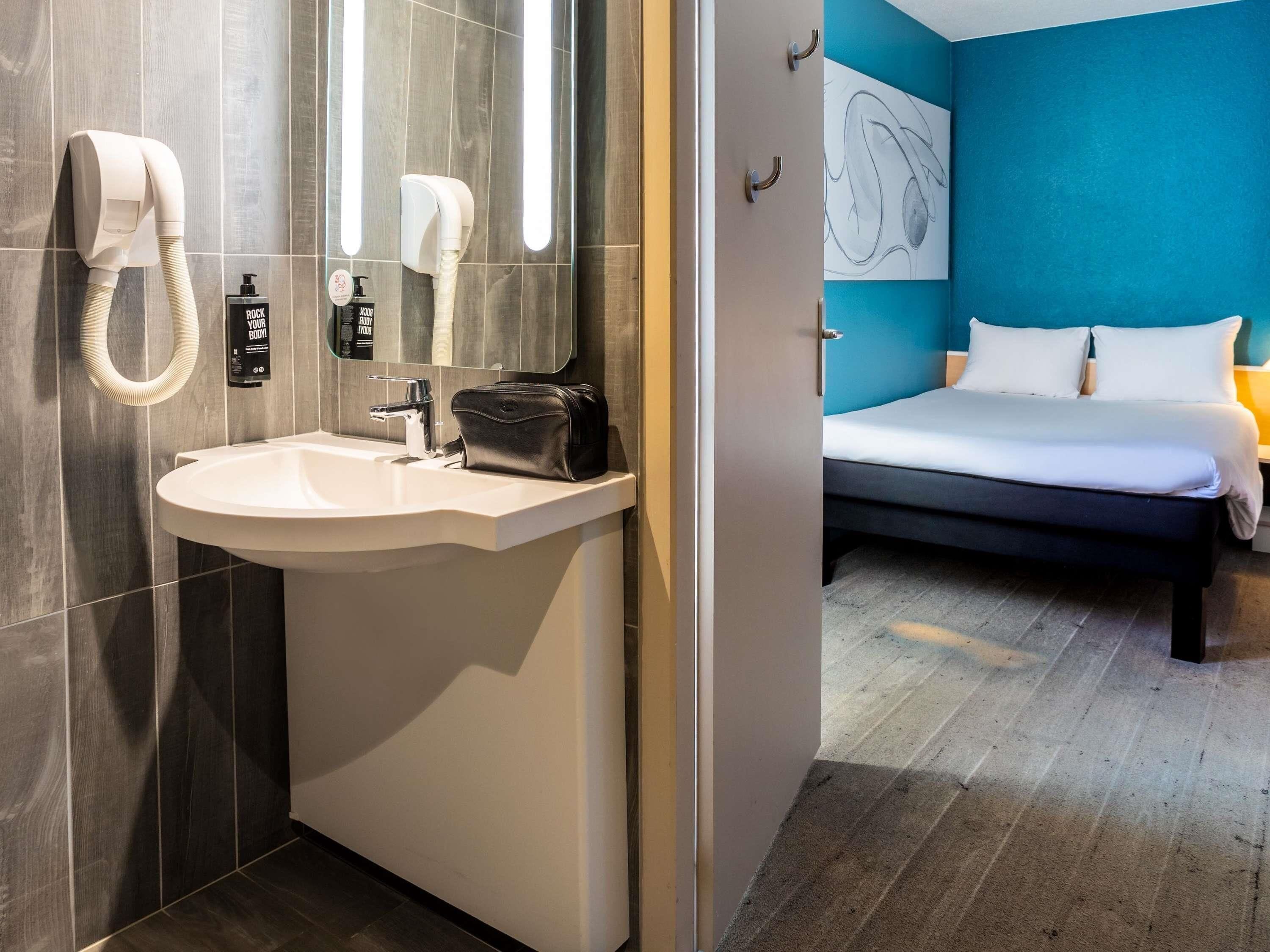 Un baño moderno con lavamanos, dispensador de toallas y secamanos. Junto al baño, una habitación con cama y una pared de acento azul.