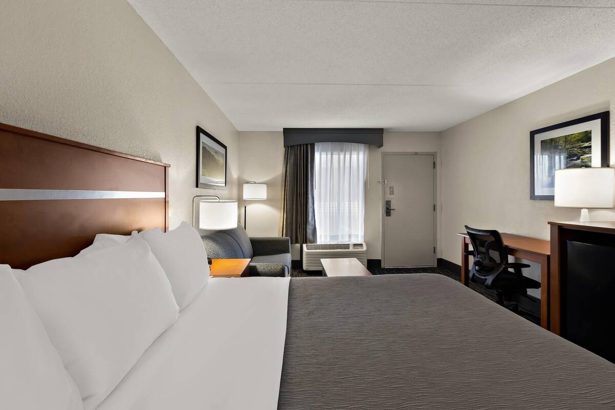 A hotel room with a large bed, a desk, a chair, a lamp, and a window with curtains.