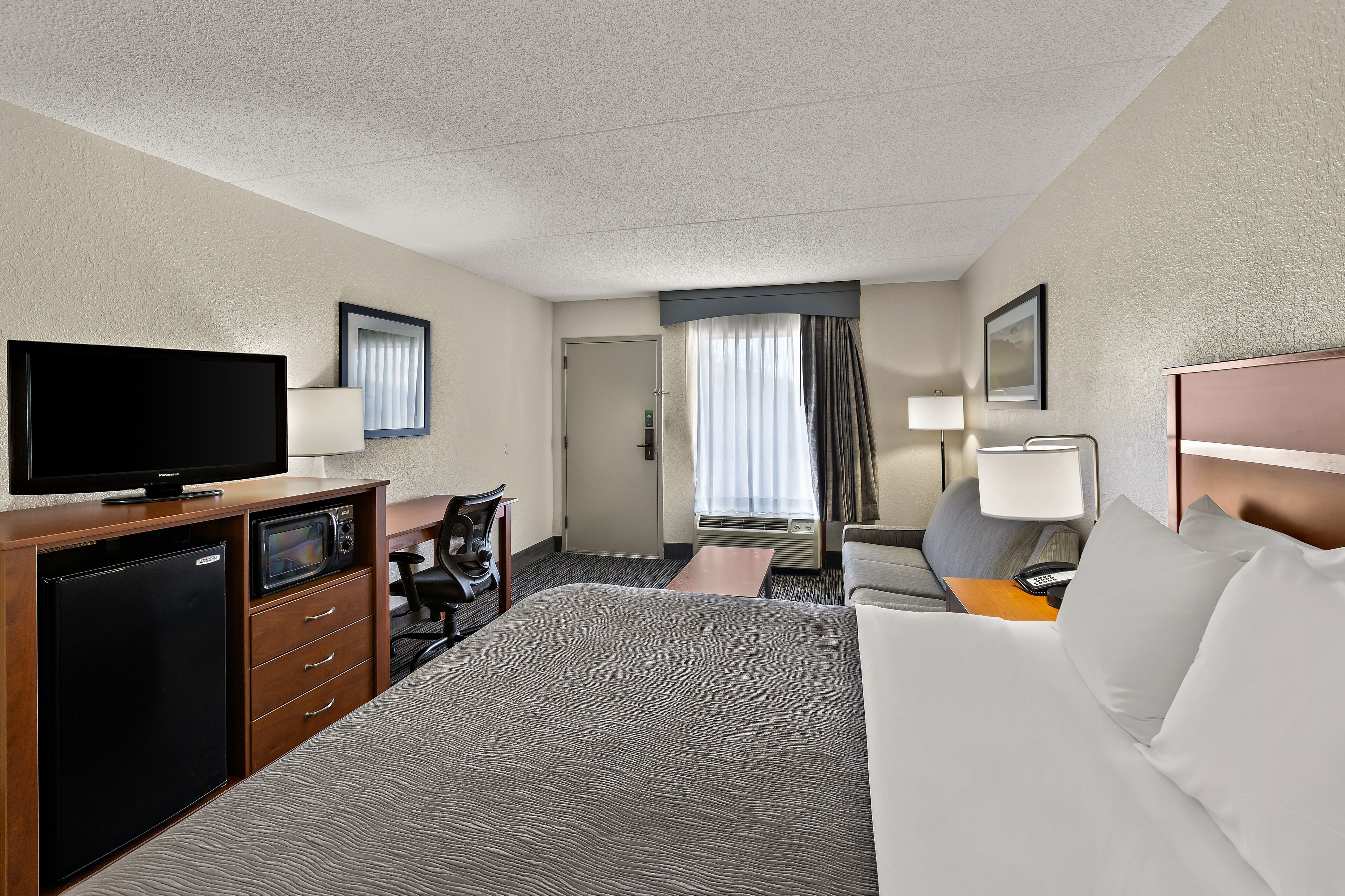 Habitación de hotel con cama, escritorio con computadora, televisión, refrigerador pequeño y ventana con cortinas.