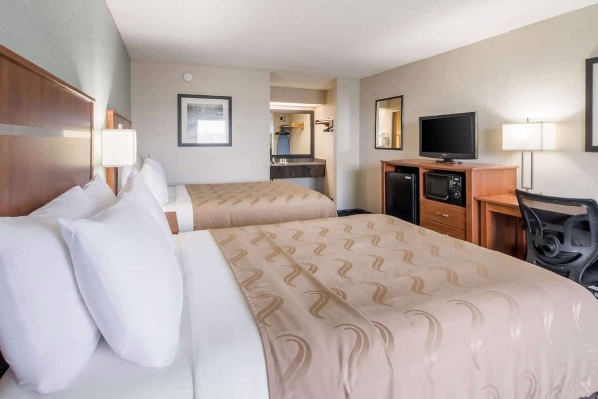 A hotel room with two beds, a desk with a television, a mirror, and a small kitchenette.