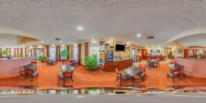 A panoramic view of a hotel lobby with a curved reception desk, multiple seating areas, and a large television screen.