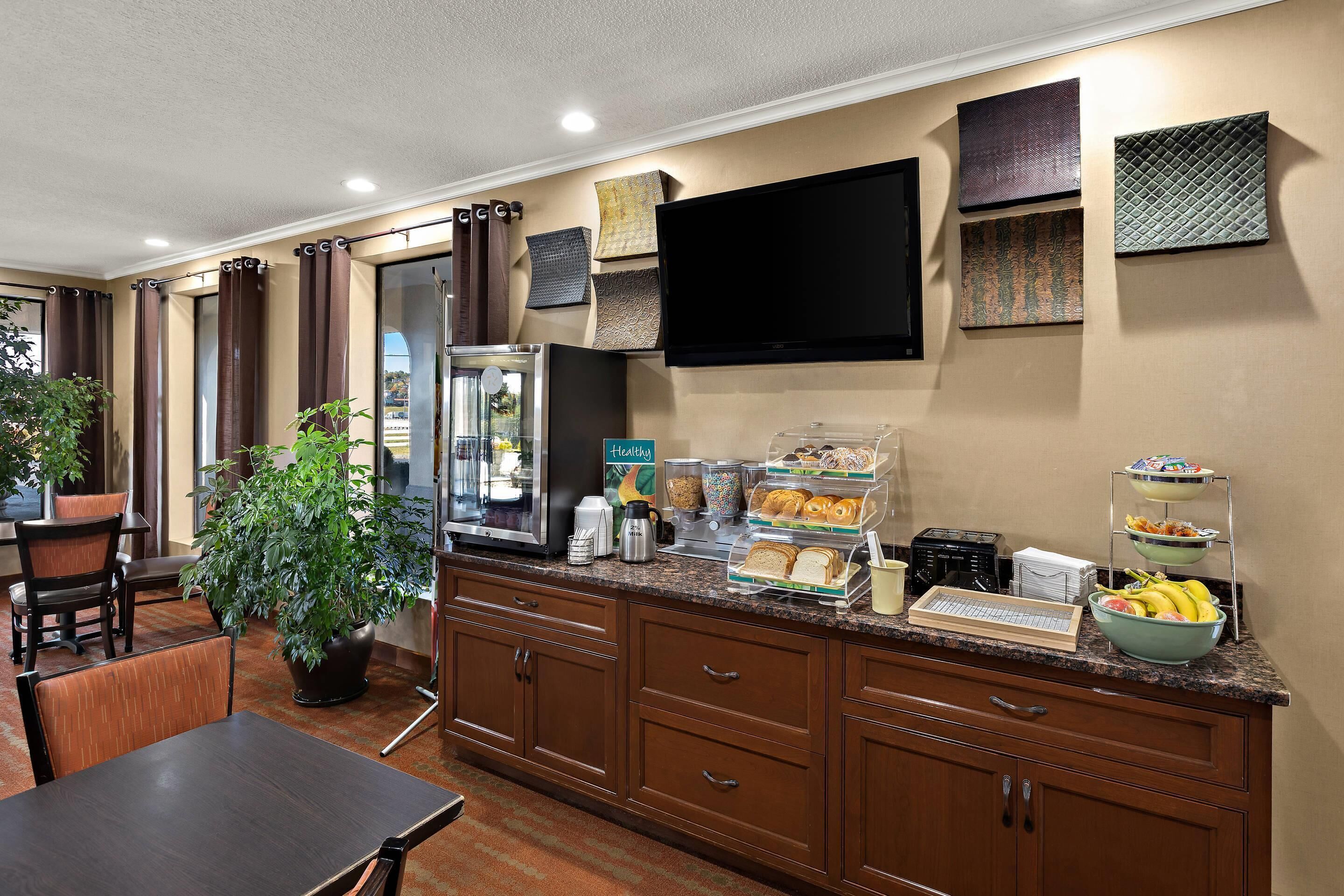 A hotel room with a kitchenette, breakfast bar, seating area, and a flat-screen TV mounted on the wall.
