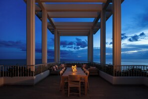 Un área de comedor al aire libre con vista al mar, que cuenta con una mesa larga y varias sillas, rodeada por una barandilla.