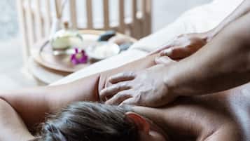 Une personne reçoit un massage dans une pièce où se trouvent une chaise en bois et un plateau avec une bouteille et une fleur.