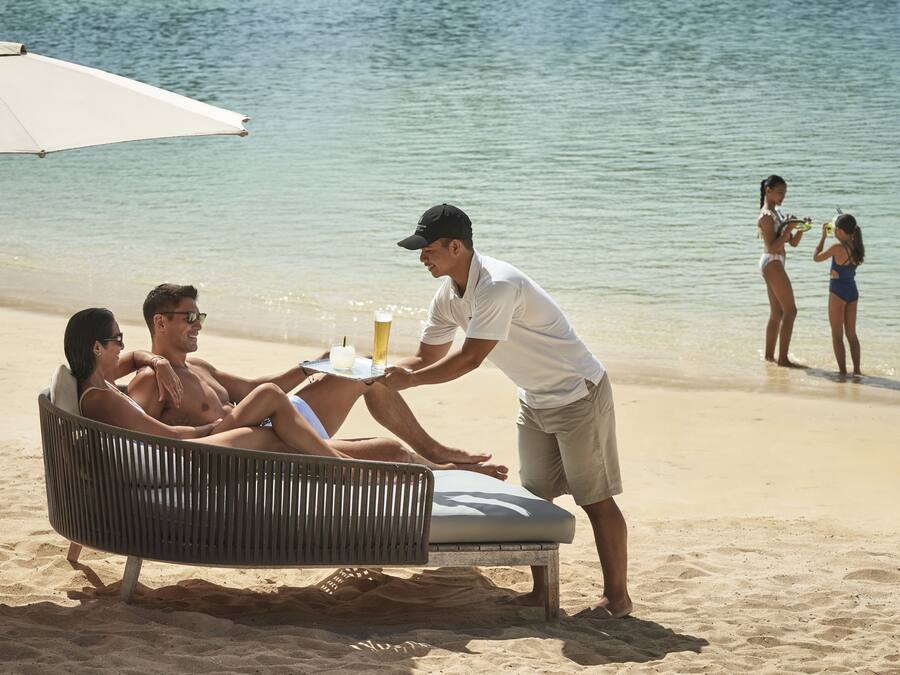 Cena de praia com um casal relaxando numa cadeira sob um guarda-sol, um garçom servindo uma bebida e uma mulher de fato de banho ao fundo.