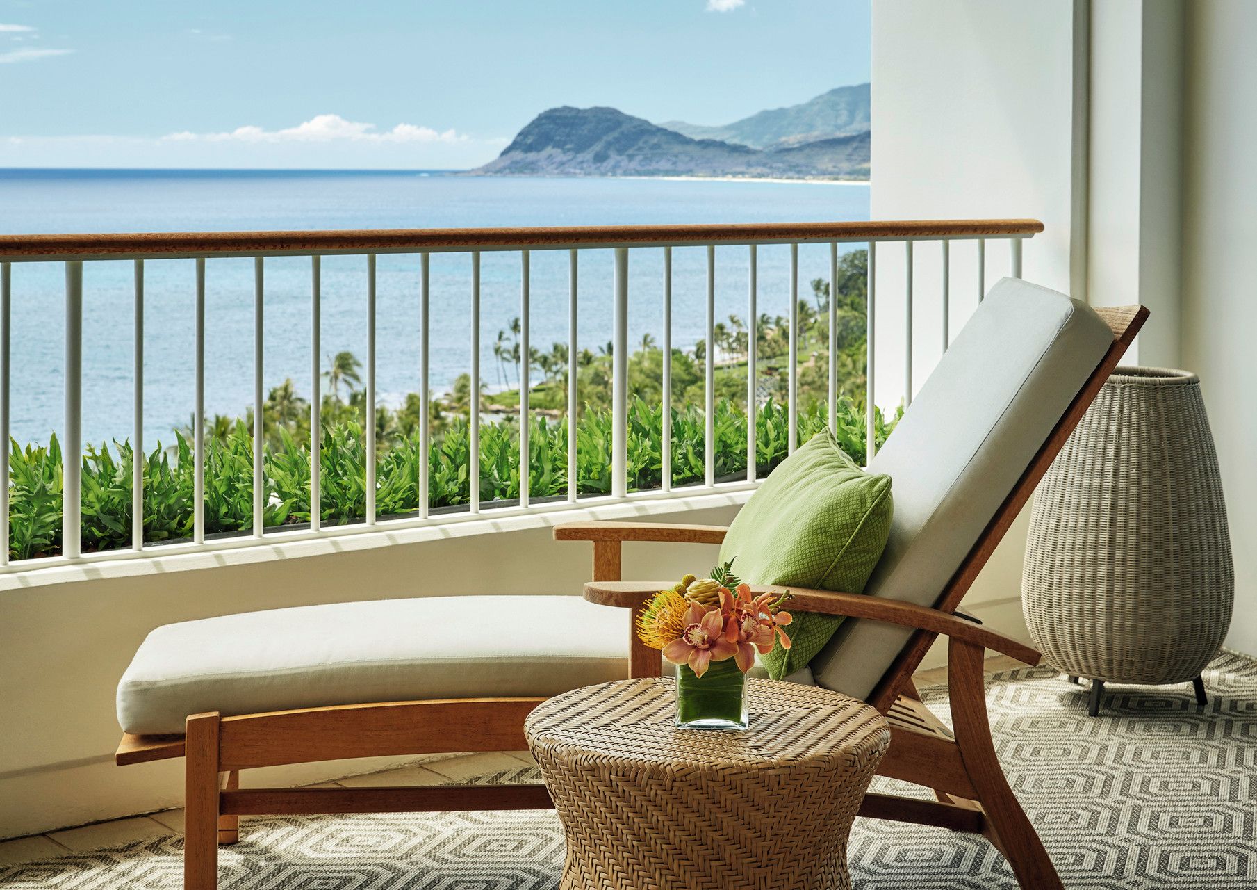 Un balcon avec vue sur l’océan et les montagnes, une chaise en osier avec un coussin et un vase rempli de fleurs.