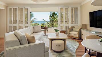 A spacious living room with a large sofa, a coffee table, and a TV. There is a balcony with a view of the ocean and palm trees.