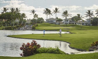 En golfbana med ett vattenhinder, palmer och bostadshus i bakgrunden.