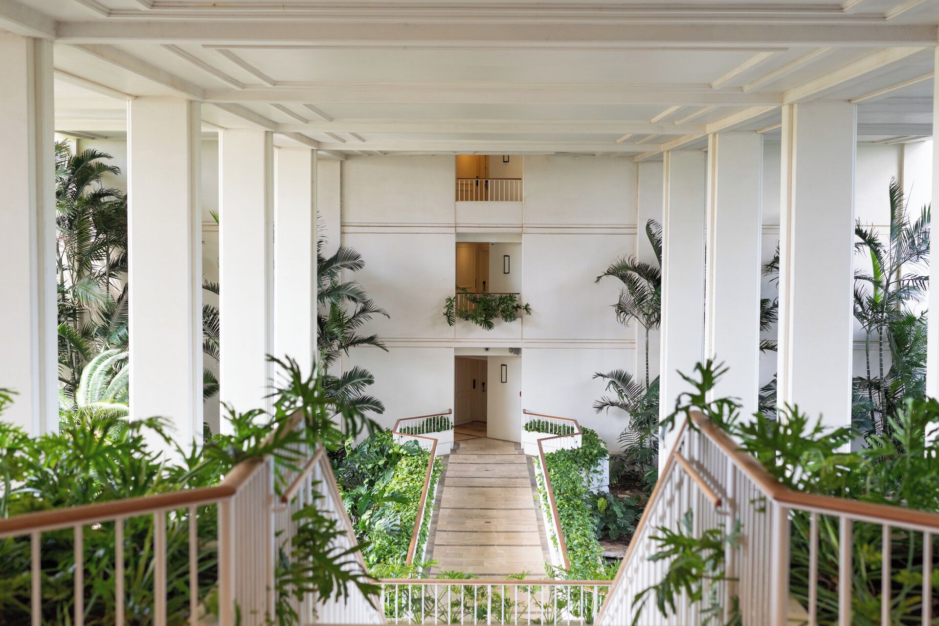 Un magnifique escalier à balustrades blanches, bordé d’une végétation luxuriante et de palmiers, mène à un palier situé au deuxième étage.