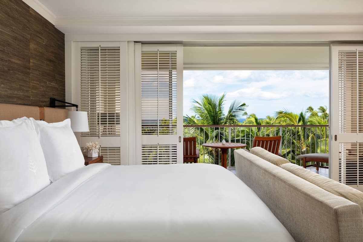 Ein Hotelzimmer mit einem großen Bett, einer Sitzecke, einem Tisch und einem Balkon mit Blick auf Palmen und den Himmel.
