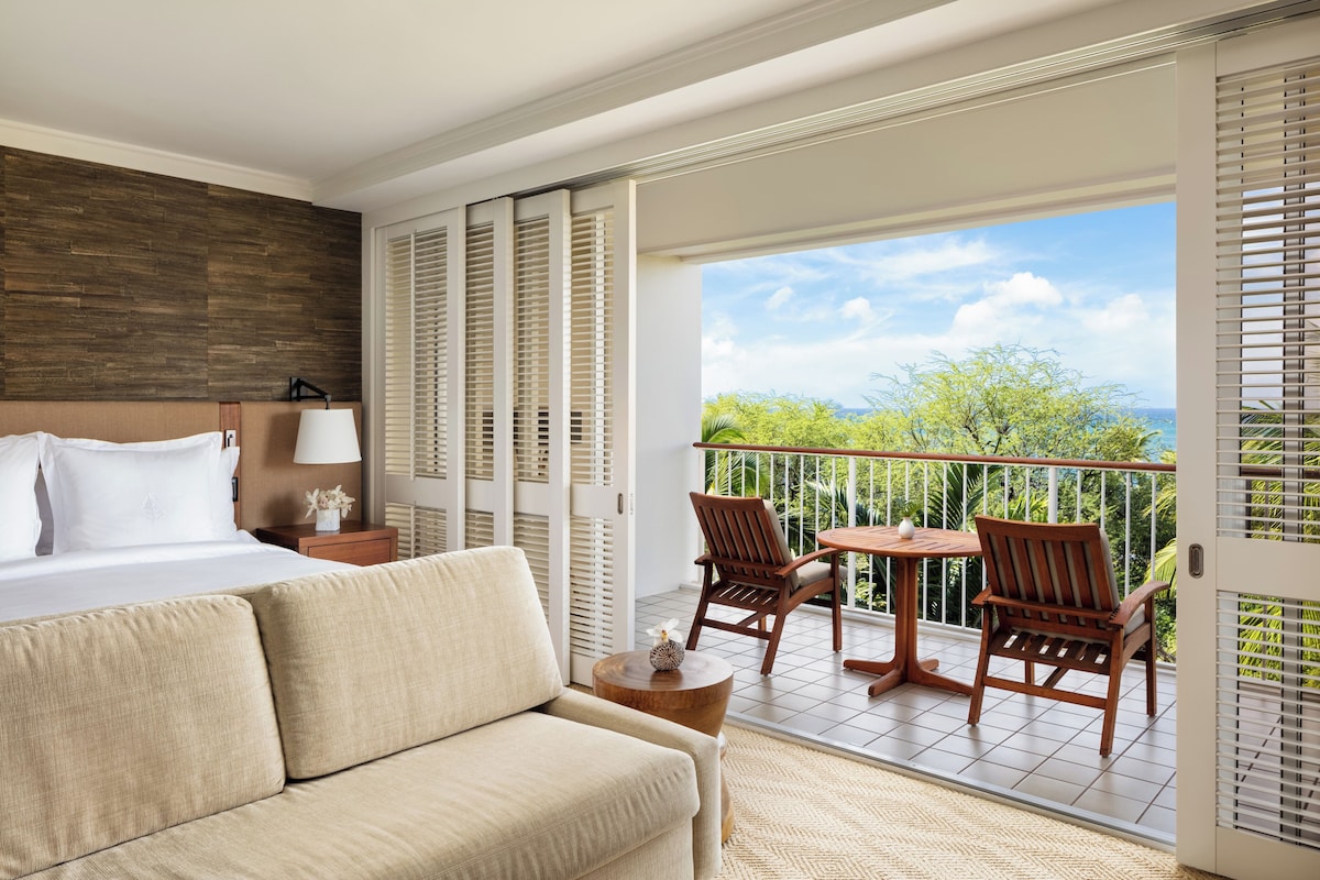 Una habitación de hotel con cama, sofá, balcón con mesa y sillas, y vista a zonas verdes y al cielo.
