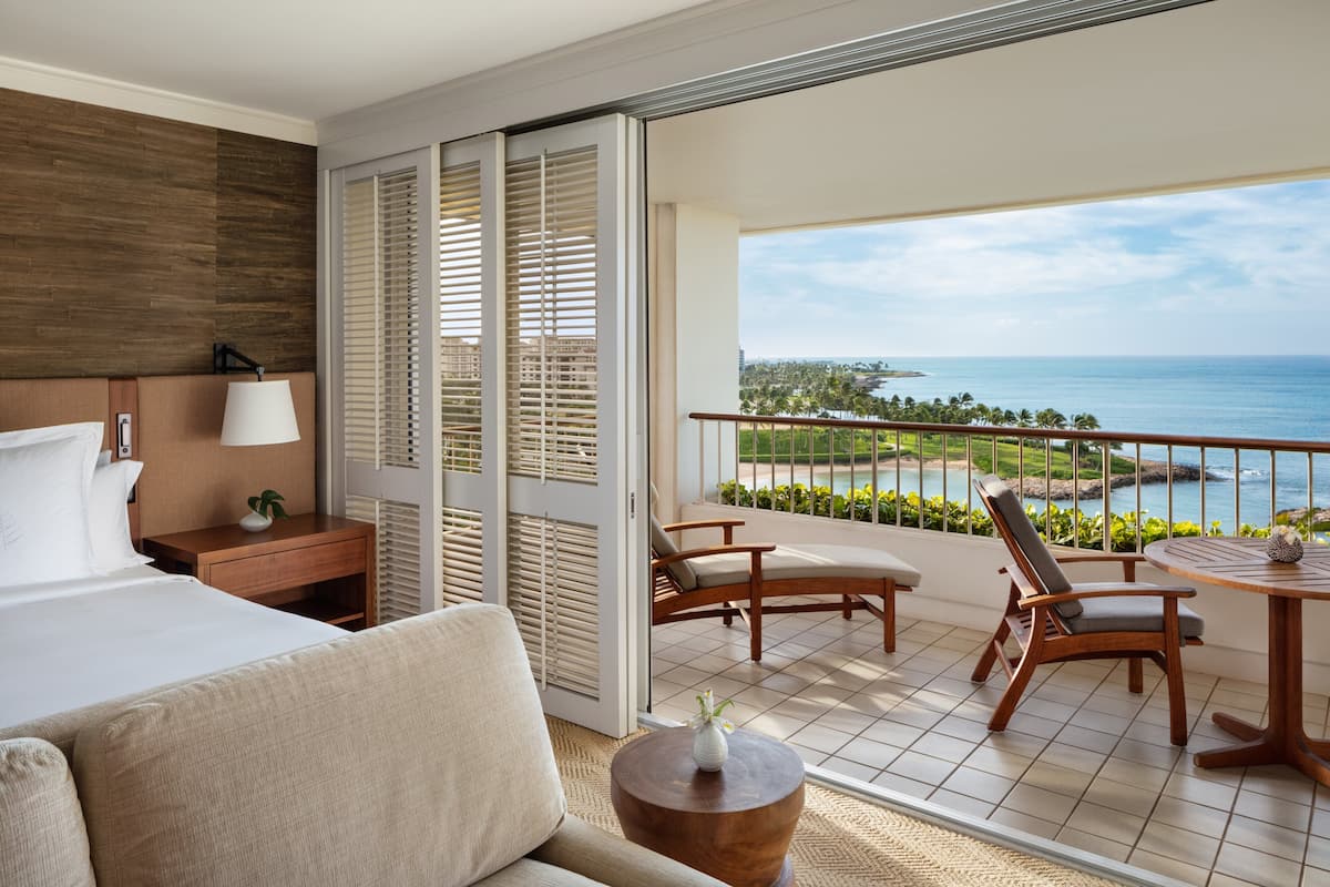 Ein Hotelzimmer mit Bett, einer Couch, einem Nachttisch aus Holz, einer Lampe, einem kleinen Tisch mit einer Vase und einem Balkon mit Blick auf das Meer und Palmen.