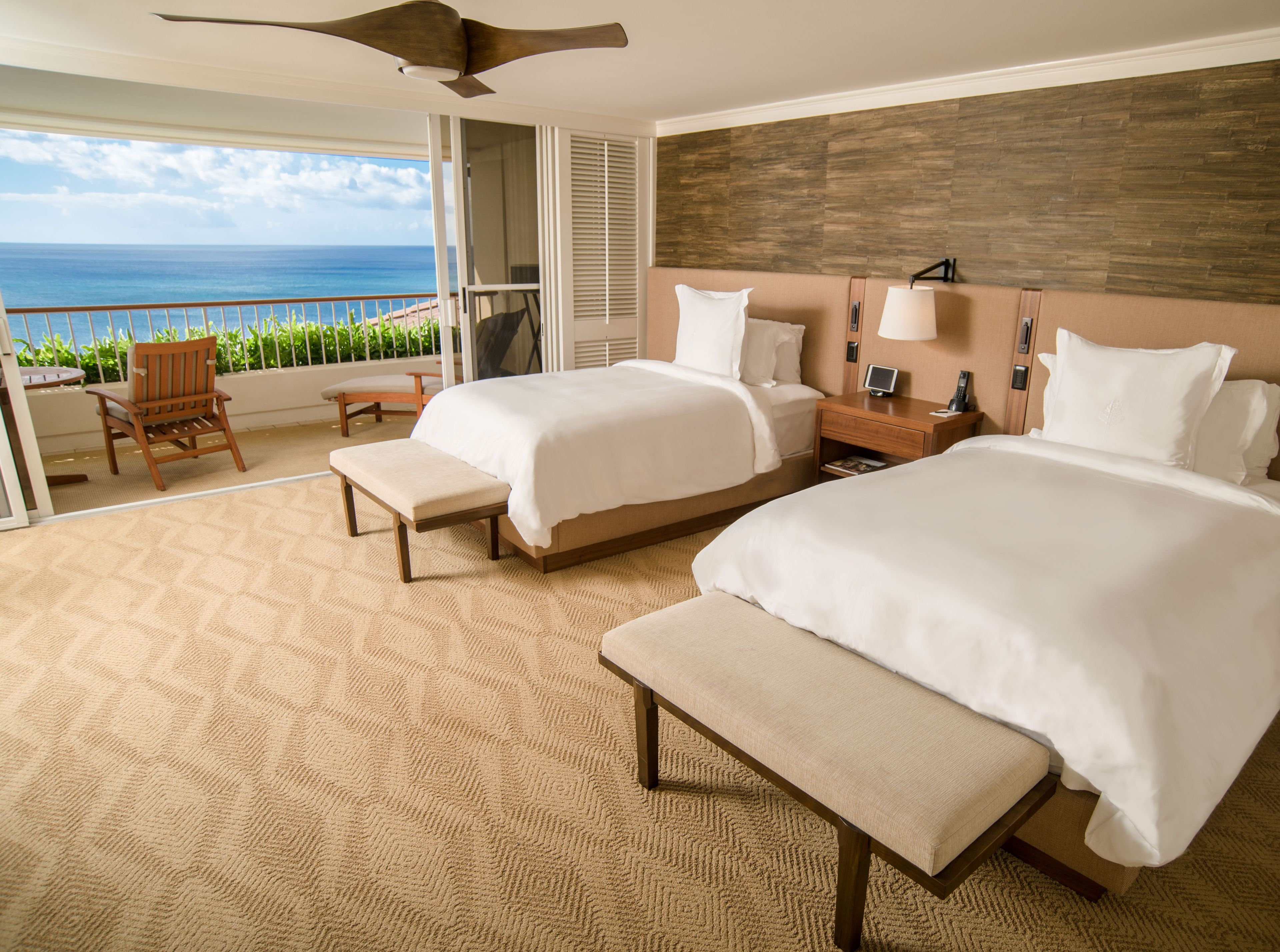 A hotel room with two beds, a bench, a chair, a nightstand, and a view of the ocean.