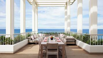 A covered outdoor seating area with a view of the ocean, featuring a table and chairs, and a white railing.