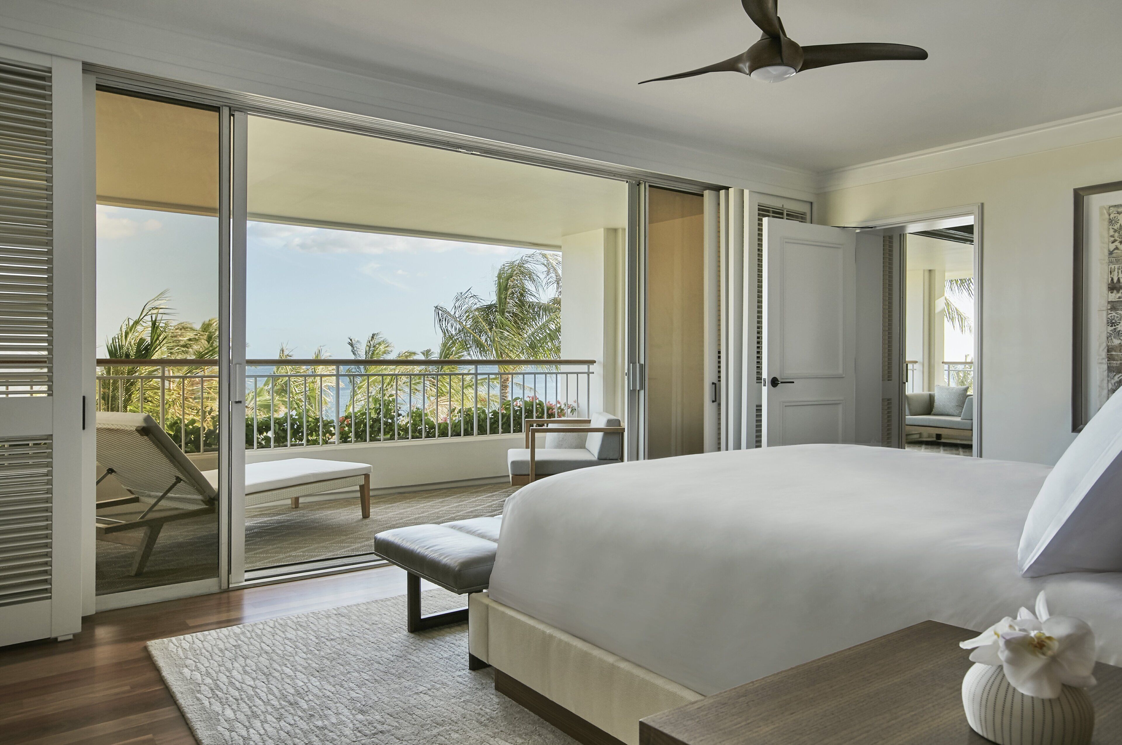 Un dormitorio con una cama grande, un balcón con vistas a palmeras y un ventilador de techo.