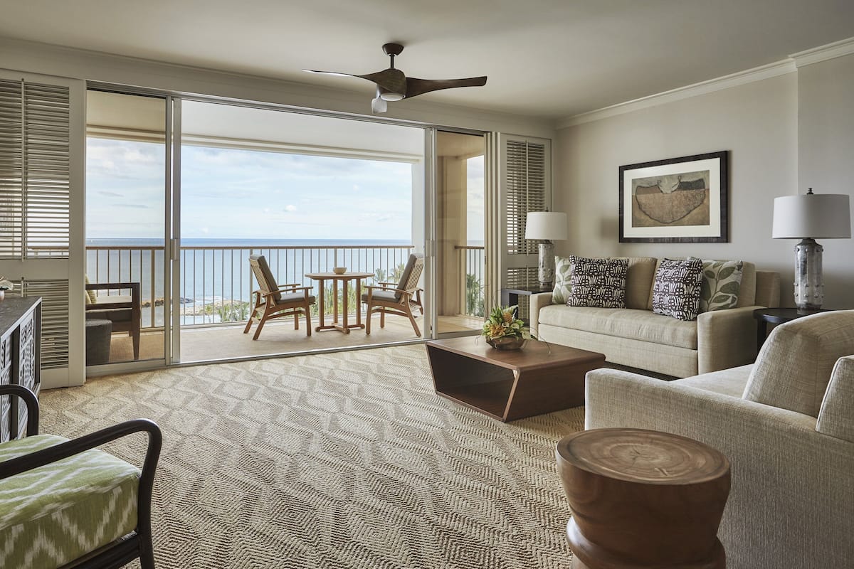 Ein geräumiges Wohnzimmer mit einer großen Schiebeglasdoor, die auf einen Balkon mit Meerblick führt. Es gibt einen Deckenventilator, ein Sofa, einen Couchtisch und eine Lampe.