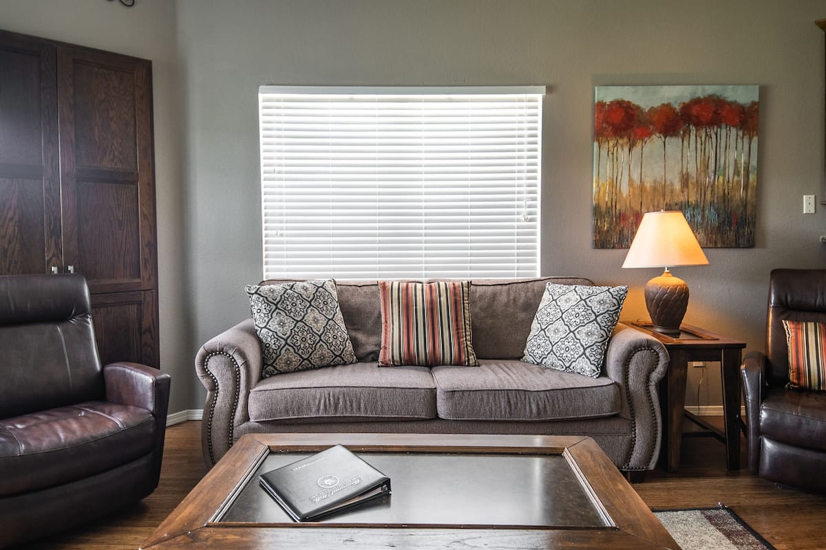 A living room with a gray sofa, two patterned pillows, a wooden coffee table with a laptop, a lamp, and a painting on the wall.