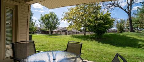 A covered patio with a round glass table and four chairs, overlooking a grassy yard with trees and houses in the background.