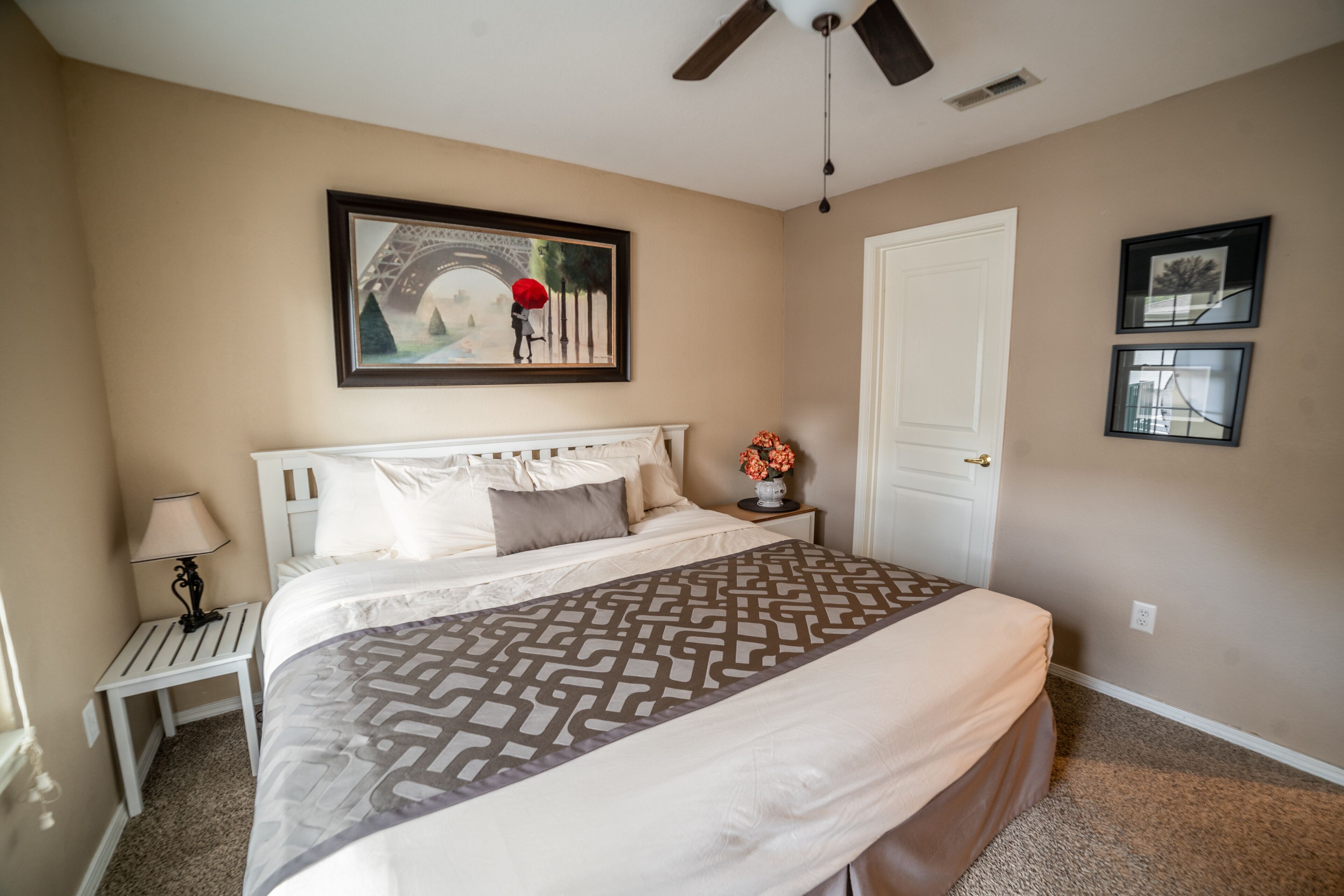 A bedroom with a bed, a nightstand, a lamp, a ceiling fan, and a door.