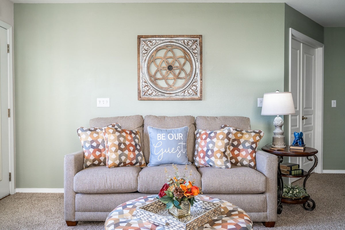 A living room with a sofa, patterned cushions, a decorative wall piece, a small side table with a lamp, and a coffee table with a floral arrangement.