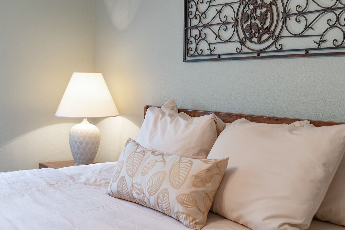 A neatly made bed with beige bedding and leaf-patterned pillows, a bedside lamp, and a decorative metal wall piece.