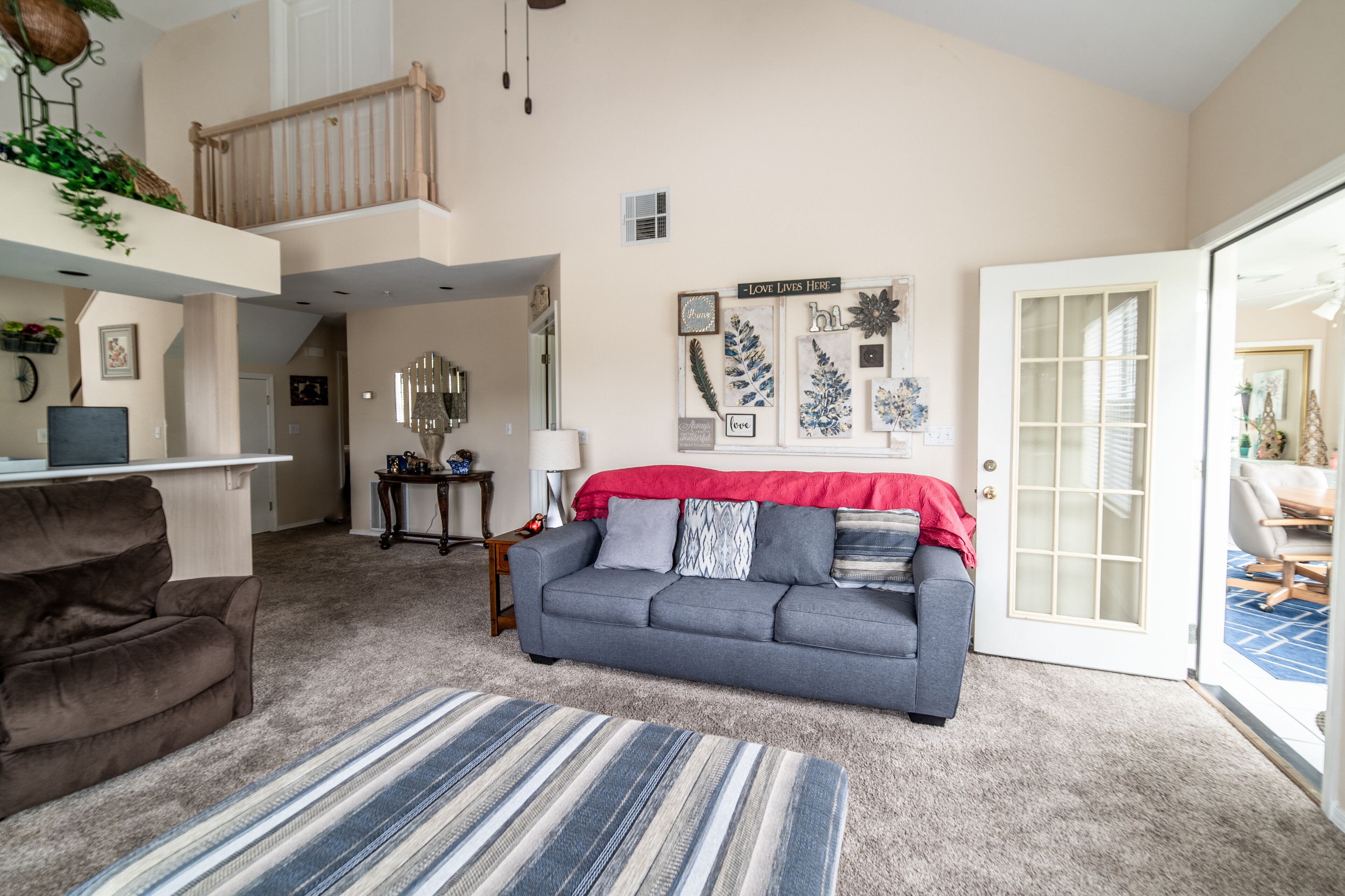 Un vaste espace de vie avec un canapé gris, un tapis à rayures et un plaid rouge. Un escalier mène à l’étage supérieur, et une porte donne accès à un balcon.