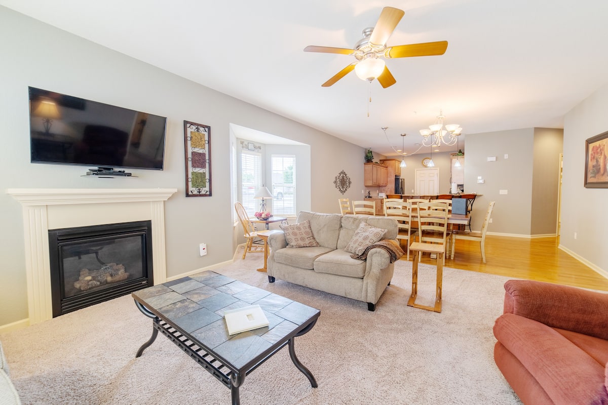 A living room with a fireplace, a television, a ceiling fan, and a dining area with a kitchen in the background.