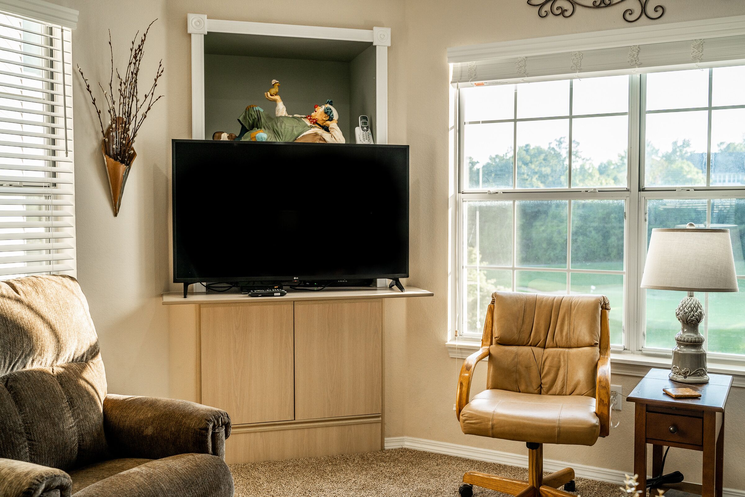 A living room with a flat-screen TV mounted on a wooden cabinet, a leather armchair, a small table with a lamp, and a window with a view of greenery.