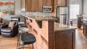 A kitchen with wooden cabinets, a stainless steel refrigerator, a microwave, and a granite countertop. There are bar stools and a leather chair. - Thousand Hills Golf Resort (Branson)