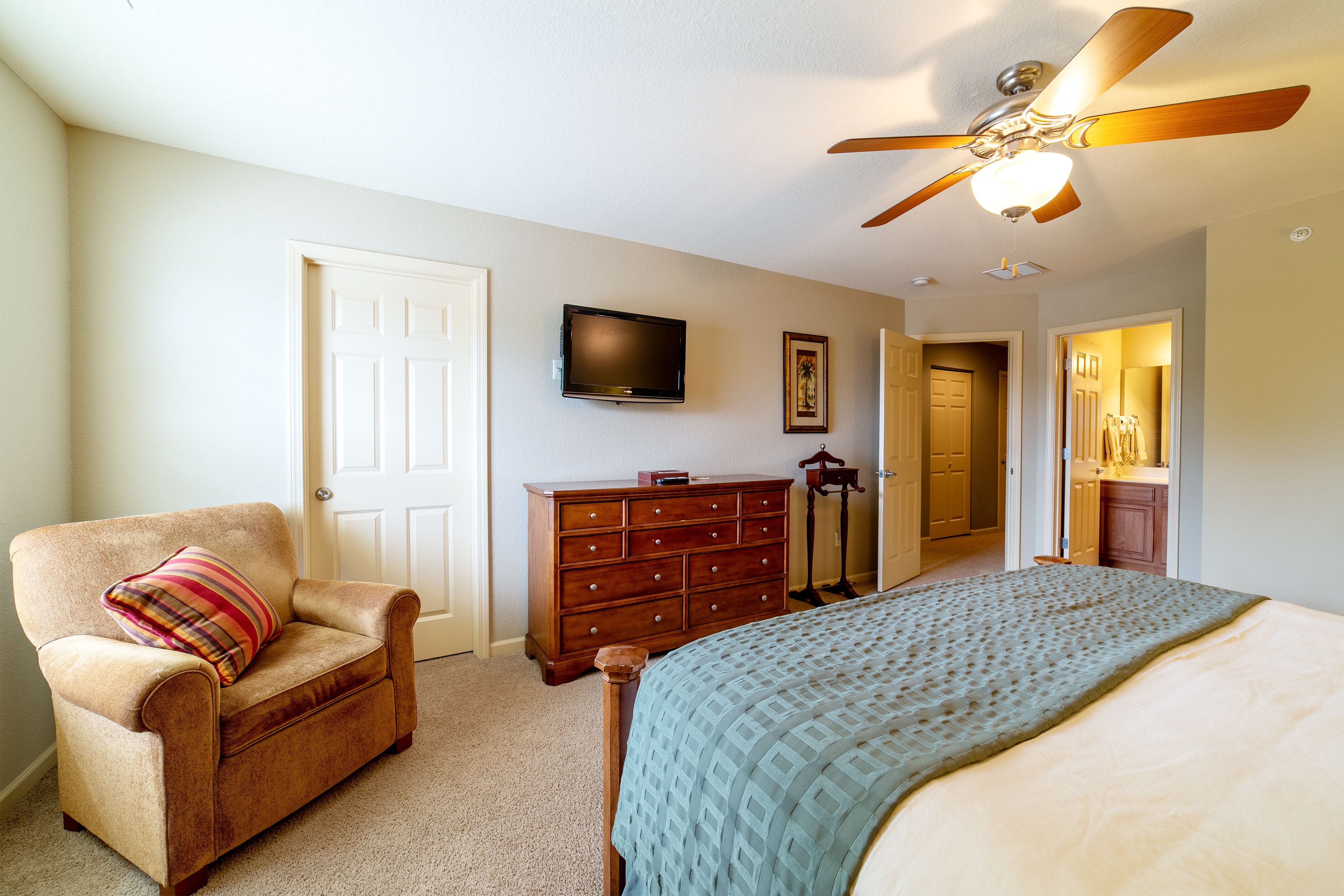 A bedroom with a bed, a chair, a dresser, a ceiling fan, and a television on the wall.