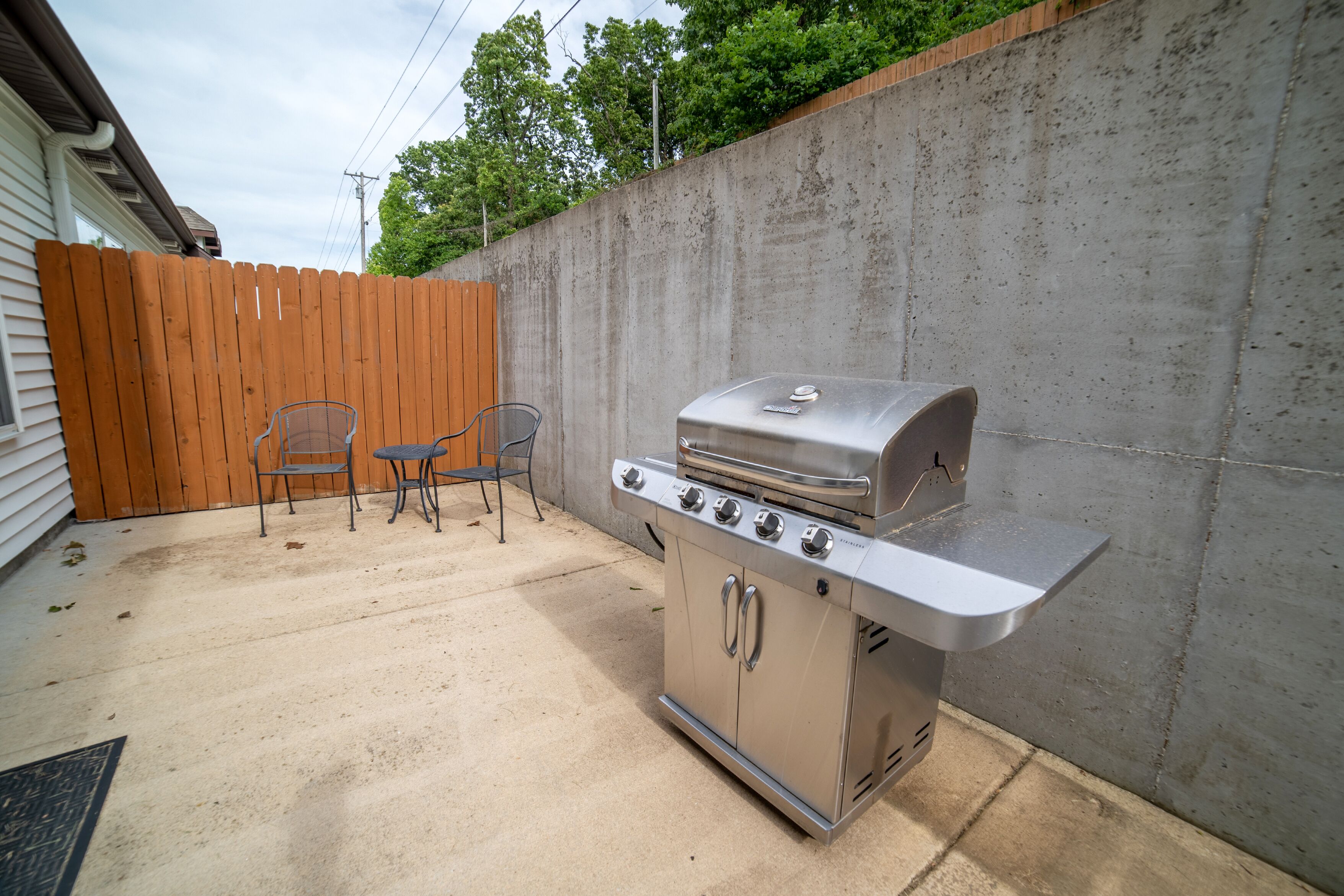 Een achtertuin met een roestvrijstalen barbecue, twee metalen stoelen en een houten schutting.