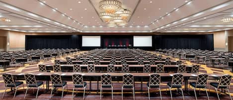 Un salón de conferencias amplio con filas de mesas y sillas, un escenario con una pantalla grande y una araña.