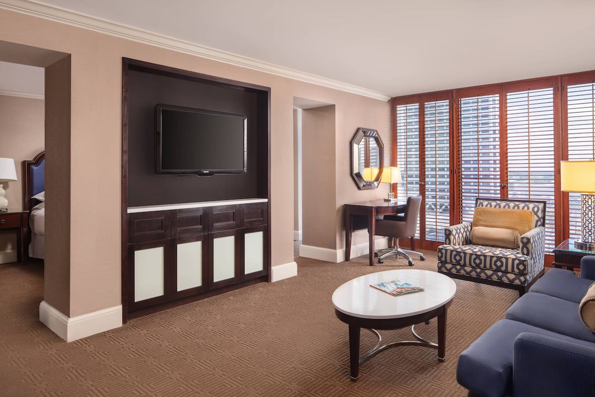 A hotel room with a large flat-screen TV mounted on a dark wooden cabinet, a round coffee table, a patterned armchair, and a desk with a lamp.