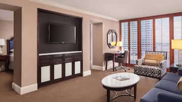 A hotel room with a large flat-screen TV mounted on a dark wooden cabinet, a round coffee table, a patterned armchair, and a desk with a lamp.