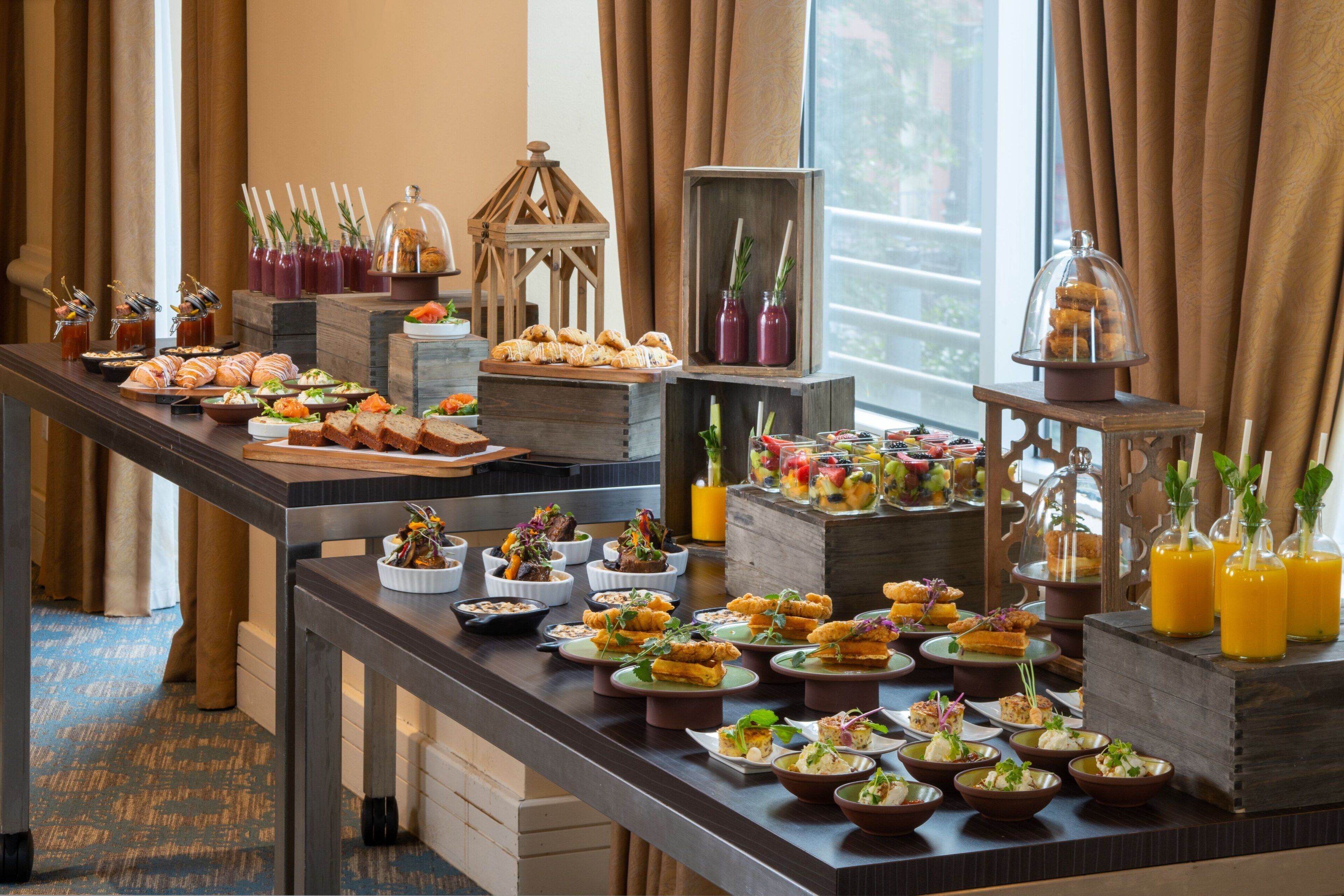 A buffet table with various food items and drinks, set against a backdrop of curtains and a window.