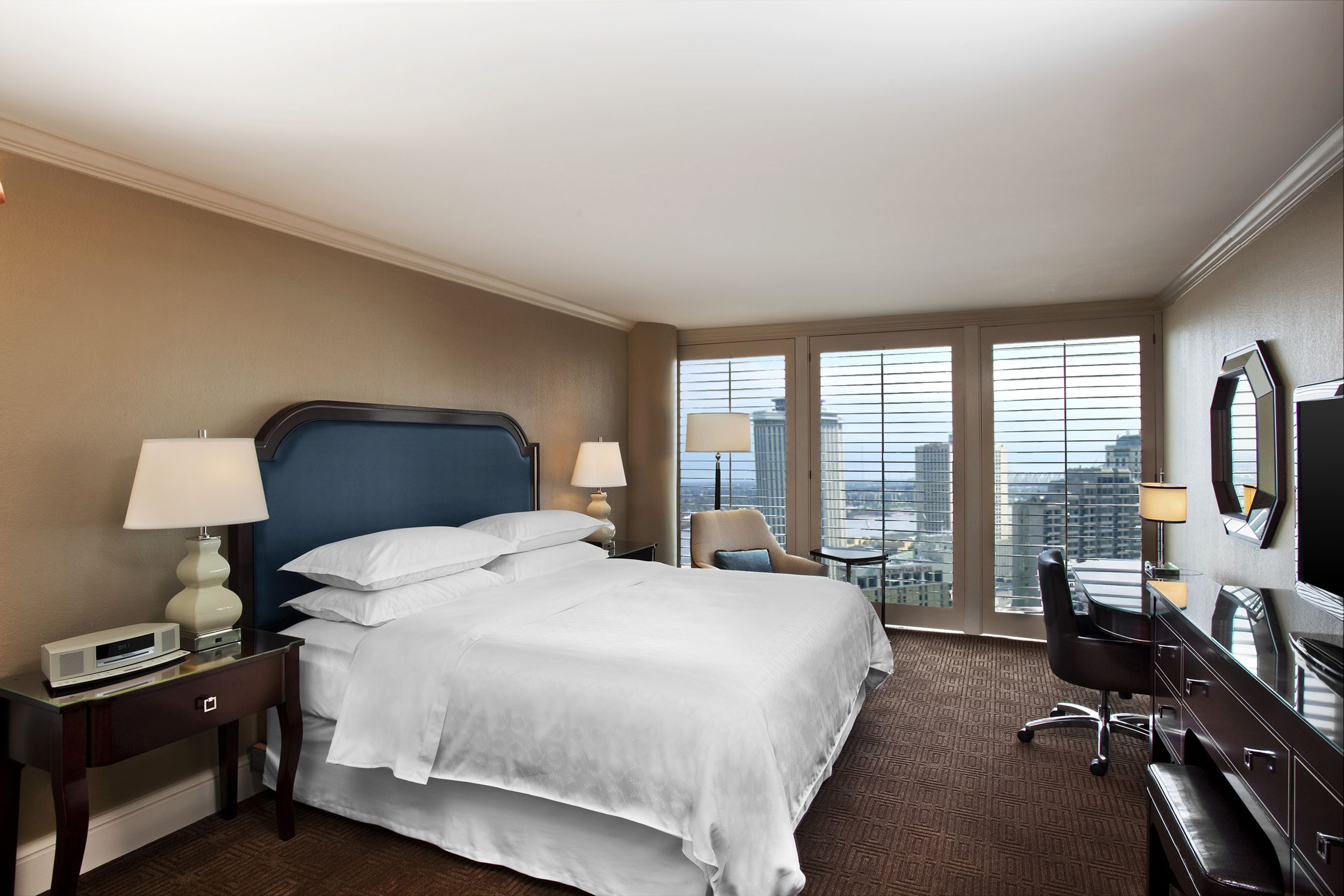 A hotel room with a large bed, two bedside tables with lamps, a desk with a chair, a TV, and a city view through large windows.